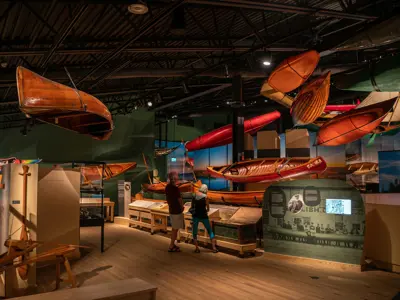 Exhibit Hall in the Canadian Canoe Museum, two people get a closer look at floor exhibit with canoes hanging from the ceiling and ehibi