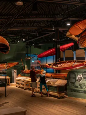 Exhibit Hall in the Canadian Canoe Museum, two people get a closer look at floor exhibit with canoes hanging from the ceiling and ehibi